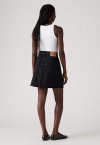 Falda de mezclilla negra con forma acampanada, de longitud hasta mitad del muslo, cinco bolsillos y un parche trasero de cuero beige. Combinada con una camiseta sin mangas blanca y zapatos planos negros.