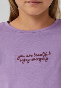 Fliederfarbenes T-Shirt mit Rundhalsausschnitt und kurzen Ärmeln, mit maroonfarbener gestickter Aufschrift "Du bist schön, genieße jeden Tag."