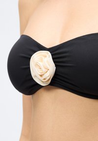 Μαύρο στράπλες μπικίνι με κεντρική λεπτομέρεια μια εκρού υφασμάτινη τριανταφυλλιά φορεμένο στο πάνω μέρος του κορμού μιας γυναίκας σε απλό φόντο.