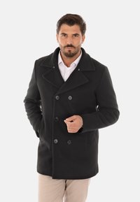 Cappotto peacoat nero in lana con design doppiopetto, bottoni grandi, rever a punta e tasche laterali, indossato sopra una camicia bianca.