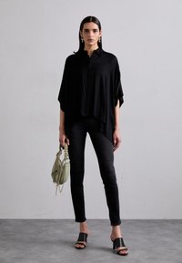 Zwarte oversized blouse met een kraag, gecombineerd met zwarte slim jeans. Zwarte slip-on sandalen en een kleine groene handtas maken de look compleet.