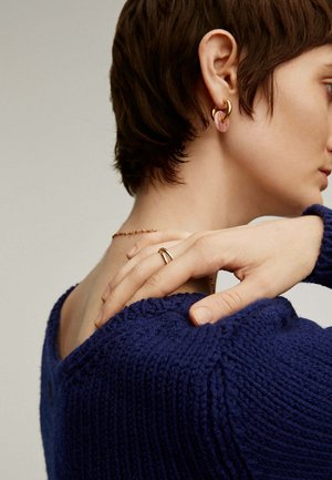 Pendientes de aro dorados y rosa, un anillo dorado en una mano y un suéter texturizado azul marino con un ancho cuello, que muestra el hombro y el cuello.