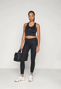 Black athletic tank top and leggings made of smooth, stretchy material. Model holds a black gym bag, wearing white sneakers.