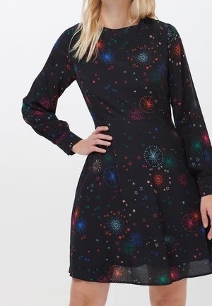 Frau trägt ein schwarzes langärmeliges Kleid mit einem bunten Muster aus Feuerwerk und Sternen, die Hand ruht auf der Hüfte.