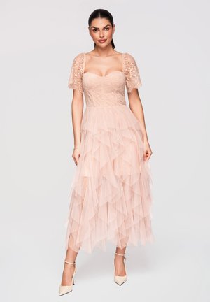 Femme debout portant une robe en dentelle rose clair avec des manches courtes bouffantes et une jupe en tulle superposée, associée à des talons crème à bride cheville.