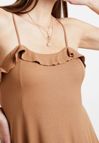Brązowy top na ramiączkach z prążkowanym wzorem, ozdobiony falbanami. Materiał jest gładki, a fason dopasowany, opada delikatnie z klatki piersiowej.