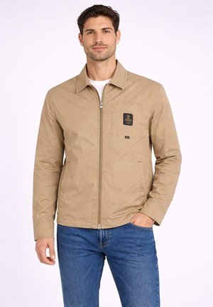 Uomo che indossa una giacca beige con zip e tasca sul petto con logo, e jeans blu, in piedi con una mano in tasca contro uno sfondo semplice.