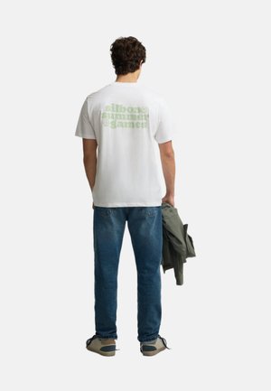 Hombre con camiseta blanca con texto en verde claro "silbon summer games" en la espalda, vaqueros azules, sosteniendo chaqueta verde en la mano derecha.