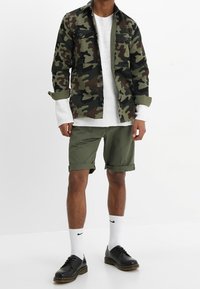 Bunda s maskáčovým vzorem v zelené a hnědé barvě, bílý pletený svetr, olivové šortky, černé boty a bílé ponožky s logem. Neformální venkovní vzhled.