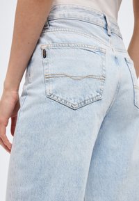 Ανοιχτό μπλε τζιν παντελόνι με ραφές στην πίσω τσέπη και μια μικρή μαύρη ετικέτα, φορεμένο από ένα άτομο με το χέρι χαλαρά δίπλα στο σώμα.