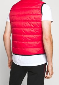 Uomo che indossa un gilet trapuntato rosso brillante sopra una camicia bianca a maniche corte e pantaloni neri, visto da dietro su uno sfondo semplice.