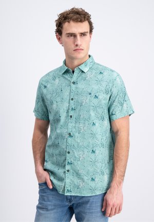 Mężczyzna w jasnoturkusowej koszuli na krótki rękaw z abstrakcyjnym kwiatowym wzorem i niebieskich dżinsach, stojący z jedną ręką w kieszeni.