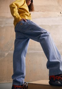 Vaqueros de mezclilla azul claro con un corte holgado, que cuentan con múltiples bolsillos y una etiqueta de marca. Combinados con una sudadera con capucha amarilla.