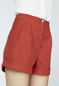 Shorts en suede rouge à taille haute, dotés de deux boutons en argent et de plis cousus. Les revers roulés ajoutent du détail à l'ourlet.