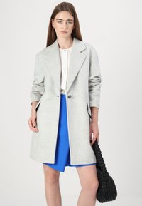 Cappotto grigio in misto lana con design sartoriale, revers a punta e maniche lunghe, abbinato a una gonna blu e una borsa nera texturizzata.