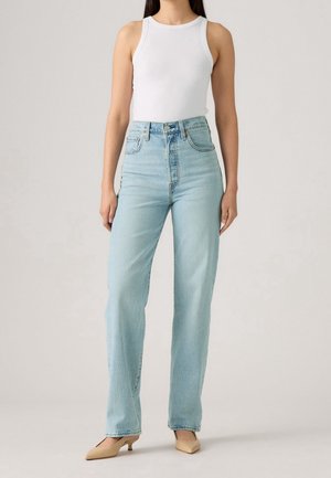 Femme portant un haut blanc sans manches, un jean bleu clair taille haute et des chaussures beiges à talons bas, debout sur un fond uni.