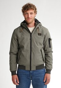 Giacca cappuccio verde oliva con zip frontale, due tasche laterali, polsini a costine e tasca con zip sulla manica. Il tessuto sembra liscio e resistente all'acqua.