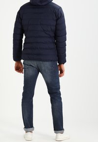 Marineblauwe pufferjack met een capuchon, met horizontale stiksels, gecombineerd met donkerblauwe denim jeans en witte sneakers.