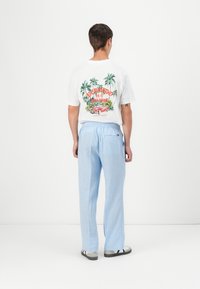 White t-shirt with colorful tropical print on back, paired with light blue, loose-fitting trousers. Casual look with elastic waistband.