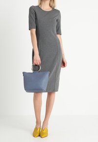 Robe-jersey grise à manches courtes ; sac à main bleu marine avec anse en anneau métallique ; ballerines pointues jaunes. Textures lisses et design ajusté.