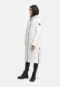 Parka bianca imbottita con design lungo, cuciture orizzontali, zip nera e tasche laterali, indossata sopra jeans scuri e stivali neri.