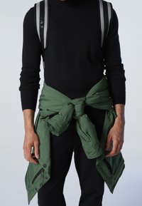 Black knitted sweater worn over an olive green puffer jacket tied at the waist. The jacket features zippers and a smooth texture. Black pants.
