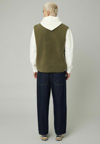 Un gilet en polaire vert olive superposé sur un hoodie blanc, associé à un pantalon en denim sombre ample et des baskets à motifs, vu de dos.