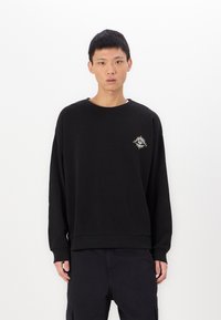 Zwarte sweatshirt met lange mouwen, ronde halslijn en een zonafbeelding in zilver op de linkerborst. Zacht materiaal met een ontspannen pasvorm.