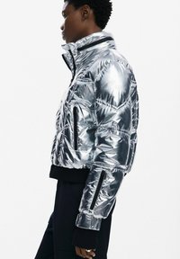 Giacca puffer argento con materiale lucido e riflettente; presenta un colletto alto, tasche con zip e polsini elasticizzati; design cropped.