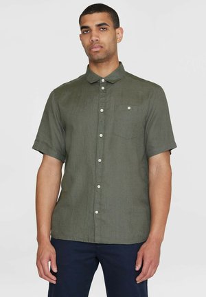 Mann trägt ein olivgrünes Hemd mit kurzen Ärmeln, Brusttasche und dunkle Hosen, steht vor einem schlichten hellen Hintergrund.