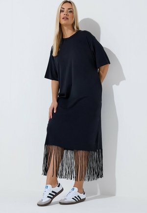 Vrouw met lang blond haar, gekleed in een zwarte midi-jurk met franjes en witte Adidas sneakers, staand tegen een witte muur.