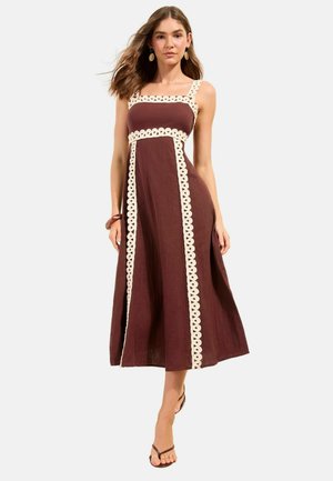 Naine, kes kannab varrukateta pruuni midi kleiti kreemika siksakääristega äärega, seisab ühe jalaga eespool, kannab kõrvarõngaid ja sandaale.