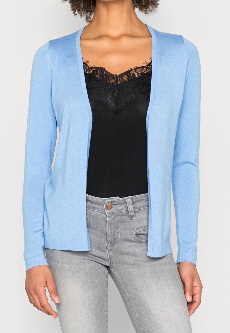 Lichtblauwe cardigan met open voorkant, lange mouwen en een zachte textuur, gelaagd over een zwart kanten tanktop en gecombineerd met grijze jeans.