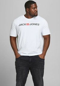 Weißes Baumwoll-T-Shirt mit einem auffälligen schwarzen Logo "JACK & JONES" auf der Vorderseite. Getragen mit dunklen Denim-Jeans im stehenden Pose.