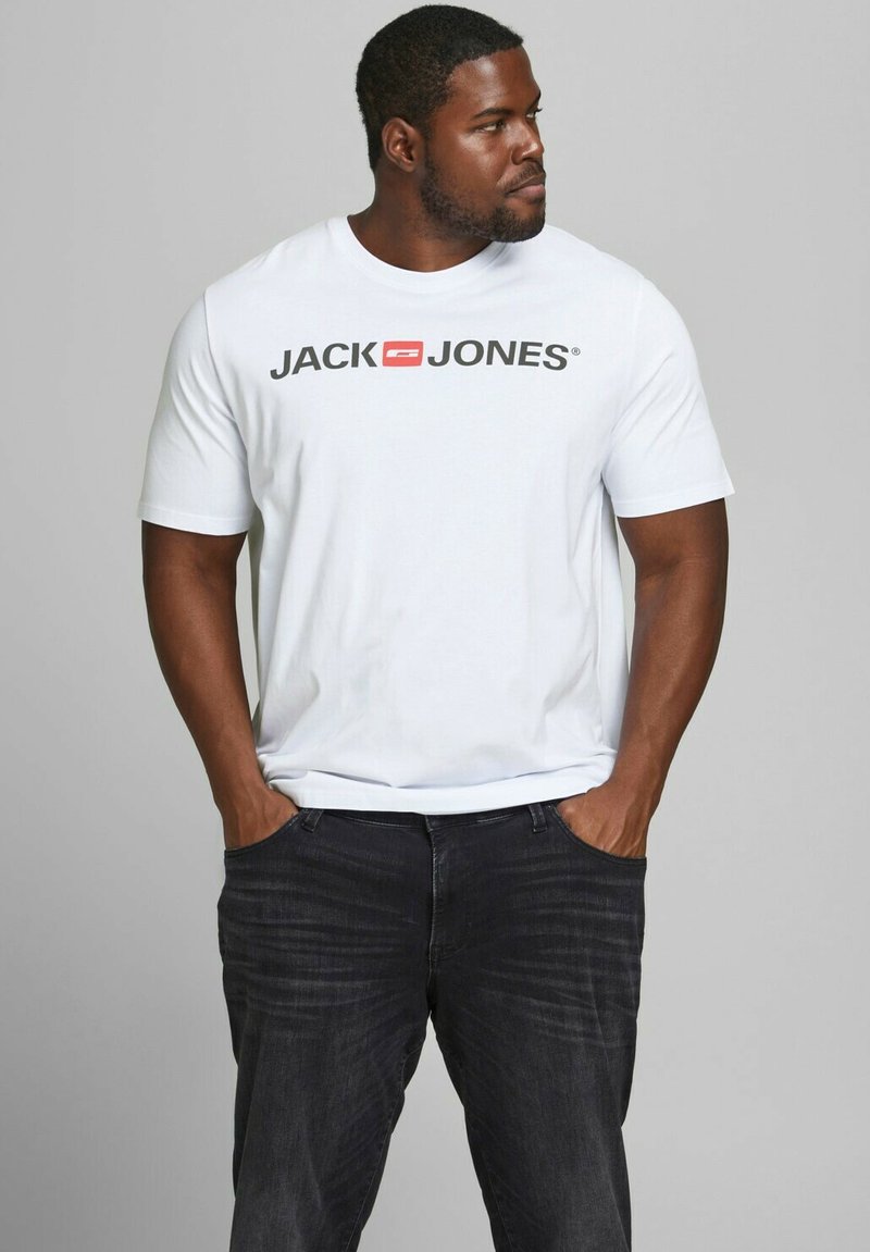 Weißes Baumwoll-T-Shirt mit einem auffälligen schwarzen Logo "JACK & JONES" auf der Vorderseite. Getragen mit dunklen Denim-Jeans im stehenden Pose.