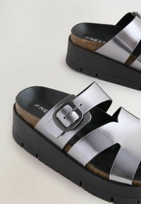 Sandali in metallo argento con design a fascia larga, accentuati da una fibbia, e una suola piatta nera testurizzata che evidenzia dettagli in sughero.