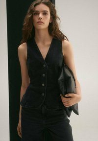 Sort denimvest med tætsiddende design, knaplukning foran og V-hals, parret med en matchende sort læderclutch. Glat overflade generelt.