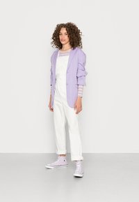 Blazer oversized de cor roxa clara, macacão branco e ténis de cano alto em lavanda. Apresenta mangas dobradas e punhos com riscas verticais.