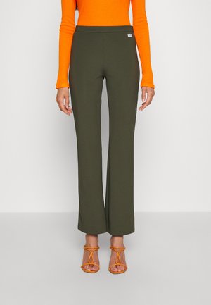 Person iført olivengrønne vide bukser, en knaldrød langærmet top og matchende orange sandaler med hæl og remme, stående mod en ensfarvet baggrund.