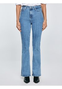 Osoba nosící světle modré džíny s vysokým pasem a rozšířenými nohavicemi a černé boty s špičatou špičkou na světlém jednobarevném pozadí.