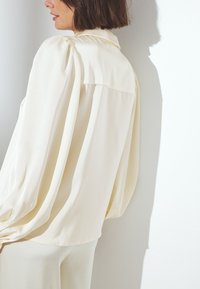 Cream-colored blouse made of smooth fabric with a relaxed fit, featuring a collar, pleated sleeves, and a rounded hem.