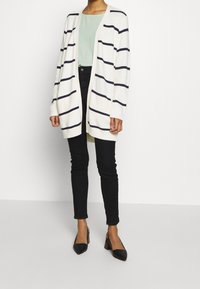 Cárdigan blanco con rayas horizontales en azul marino, mangas largas, abertura frontal, combinado con una blusa verde clara y vaqueros skinny negros, tacones negros de punta.