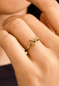 Bague en or avec un accent en forme de nœud sur le dessus, finition lisse et brillante, et une bande fine, portée sur une main avec une texture de peau visible.