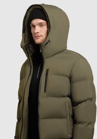 Maslinasto zeleni puffer jakna s visokim ovratnikom, velikom kapuljačom i zatvaračima na džepovima. Glatka tekstura sa plišanom, podstavljenom bojom.
