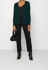 Cárdigan de punto verde con escote en V, combinado con una blusa negra con encaje, jeans negros de cintura alta y botines negros de tacón.
