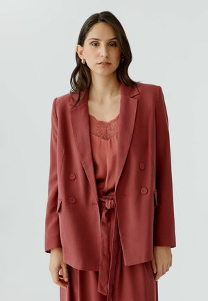 Femme aux longs cheveux foncés portant un blazer et une robe assortis de couleur rouille avec des détails en dentelle, debout devant un fond clair uni.