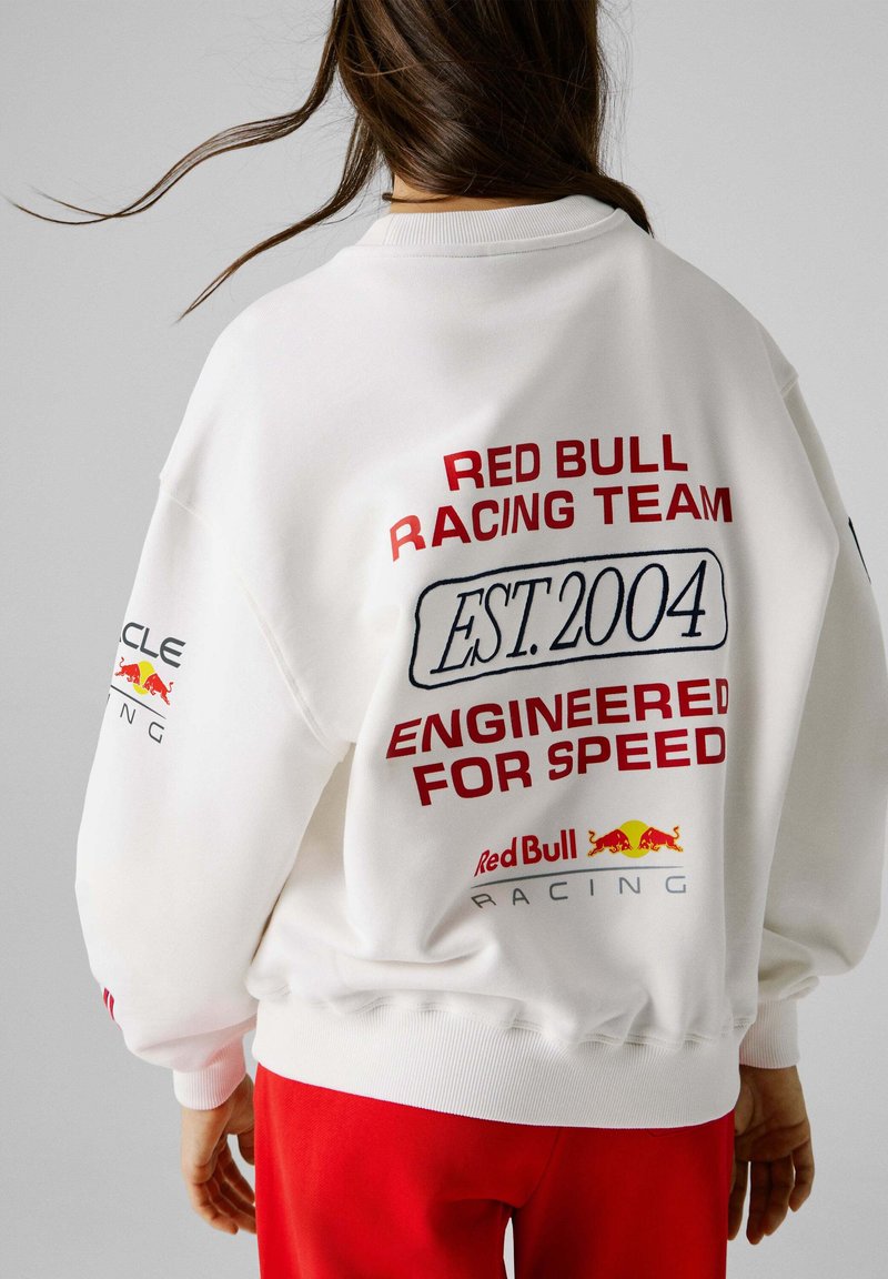 Άτομο που φοράει άσπρο φούτερ Red Bull Racing με τα κείμενα "EST. 2004" και "Engineered for Speed", συνδυασμένο με κόκκινο παντελόνι, από πίσω.