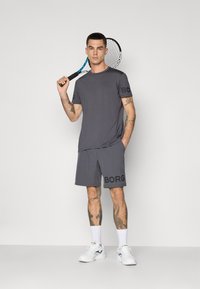 Mann in grauem Sportoutfit, der einen Tennisschläger über der Schulter hält und vor einem schlichten Hintergrund steht, mit Tattoos an den Armen und Beinen.