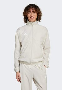 Giacca zip-up beige chiaro realizzata in tessuto liscio, con colletto alto, polsini elasticizzati e un logo riconoscibile sul petto.