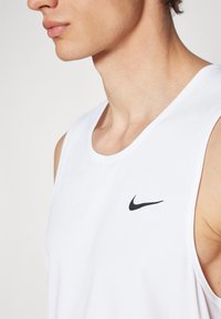 Bijela atletska majica bez rukava s okruglim izrezom, koja na gornjoj lijevoj strani ima crni Nike swoosh logo i glatku teksturu sličnu pamuku.